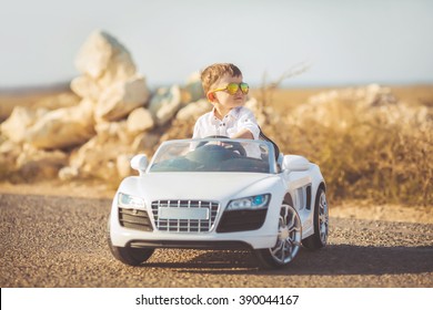 Funny boy car driver with the steering wheel. year-old boy in a white shirt in a red toy car in the street. Little boy driving big toy car and having fun, outdoors. Young kid portrait with toy car