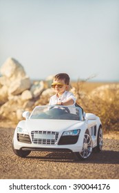 Funny Boy Car Driver With The Steering Wheel. Year-old Boy In A White Shirt In A Red Toy Car In The Street. Little Boy Driving Big Toy Car And Having Fun, Outdoors. Young Kid Portrait With Toy Car