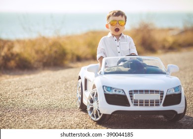 Funny boy car driver with the steering wheel. year-old boy in a white shirt in a red toy car in the street. Little boy driving big toy car and having fun, outdoors. Young kid portrait with toy car