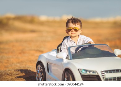 Funny Boy Car Driver With The Steering Wheel. Year-old Boy In A White Shirt In A Red Toy Car In The Street. Little Boy Driving Big Toy Car And Having Fun, Outdoors. Young Kid Portrait With Toy Car