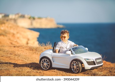 Funny Boy Car Driver With The Steering Wheel. Year-old Boy In A White Shirt In A Red Toy Car In The Street. Little Boy Driving Big Toy Car And Having Fun, Outdoors. Young Kid Portrait With Toy Car