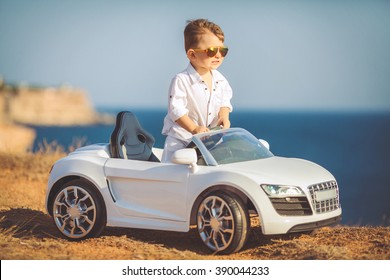 Funny Boy Car Driver With The Steering Wheel. Year-old Boy In A White Shirt In A Red Toy Car In The Street. Little Boy Driving Big Toy Car And Having Fun, Outdoors. Young Kid Portrait With Toy Car