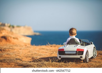 Funny Boy Car Driver With The Steering Wheel. Year-old Boy In A White Shirt In A Red Toy Car In The Street. Little Boy Driving Big Toy Car And Having Fun, Outdoors. Young Kid Portrait With Toy Car