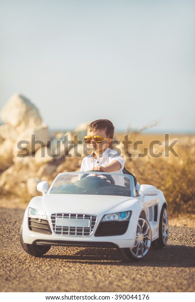 Funny boy car driver with the steering wheel.\
year-old boy in a white shirt in a red toy car in the street.\
Little boy driving big toy car and having fun, outdoors. Young kid\
portrait with toy car