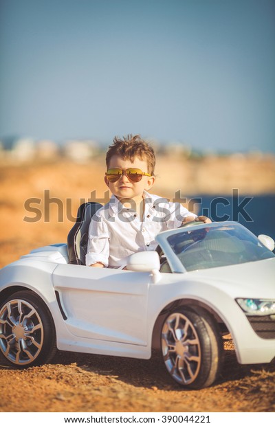 Funny boy car driver with the steering wheel.\
year-old boy in a white shirt in a red toy car in the street.\
Little boy driving big toy car and having fun, outdoors. Young kid\
portrait with toy car