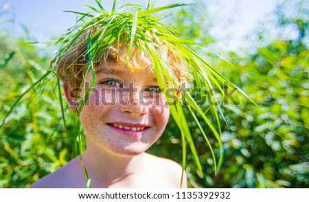 funny boy with freckles and grass on his head smiling, close-up portrait, happy young kid with green grass hair.