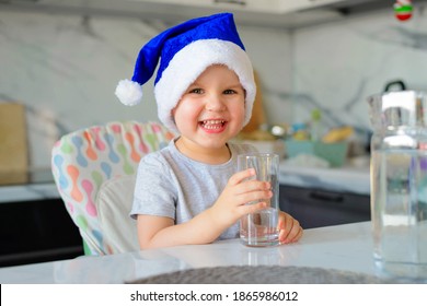 Funny Boy Kid In A Blue Santa Hat Drinking Filtered Water From A Glass In The Kitchen. Holidays, Health Concept.