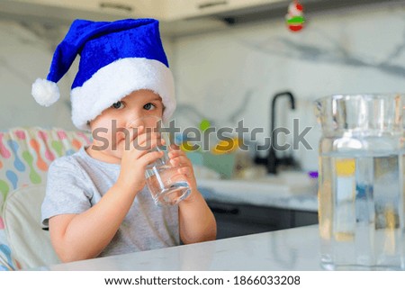 Funny boy kid in a blue Santa hat drinking filtered water from a glass in the kitchen. Holidays, health concept.