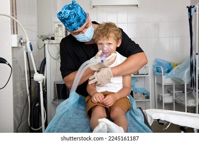 Funny boy sits on couch in hospital room, breathing narcosis through ventilation mask in hands of a professional doctor. The doctor in a colored cap pressed the boy to himself, holds an oxygen mask - Powered by Shutterstock