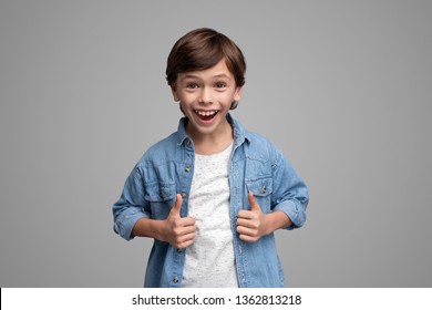 Funny boy in stylish outfit showing thumb up gesture with both hands and looking at camera with amazed face expression while standing on gray background - Shutterstock ID 1362813218