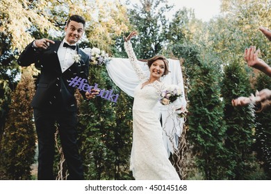 Funny Bride And Groom Jump Up Holding A Lettering With Wedding Hashtag In Their Arms