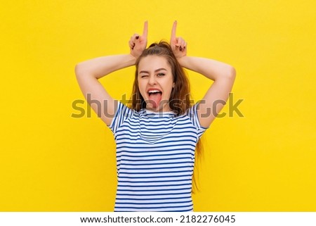 Funny brunette girl make horns by fingers, making faces, showing tongue, having fun, grimacing, fooling, teasing, standing in white-blue striped t shirt over yellow background