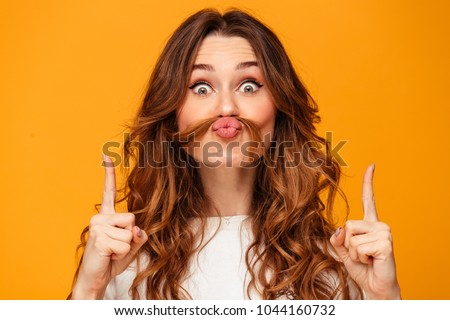 Funny brunette woman in sweater holding hair on face like mustache while pointing up and looking at the camera over yellow background