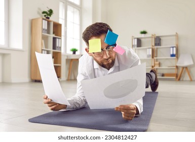 Funny busy businessman multitasking in office. Businessman with post it memo notes on his face reading financial papers while doing plank exercise on yoga mat on office floor