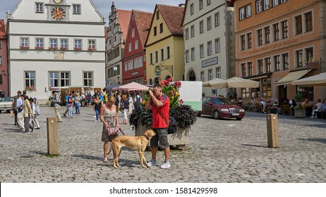 Funny, Candid Photo Of A Photographer So Busy Taking Pictures He Seems Oblivious To His Dog Taking A Good Sniff Of His Crotch.  Rothenburg, Germany. Aug 2019.                               