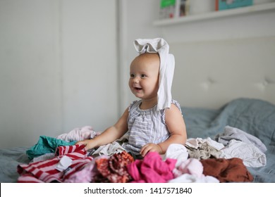 Funny Caucasian Baby 10 Months Playing With A Bunch Of Clothes, A Mess And Children, Motherhood And Mom Do Not Have Time, Rummaged At Home. Bright Bedroom Interior