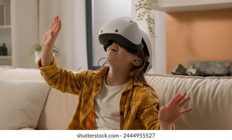 Funny caucasian kid girl child schoolgirl having fun in virtual reality headset at home couch playing online video game in world metaverse using innovative technology VR glasses exploring cyberspace
