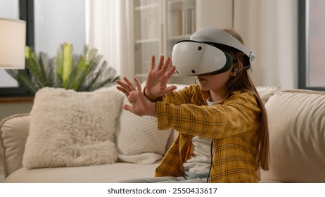 Funny caucasian kid girl child schoolgirl having fun in virtual reality headset at home couch playing online video game in world metaverse using innovative technology VR glasses exploring cyberspace