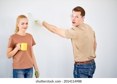 Funny Caucasian Married Couple Paint The Wall, Woman Have Rest, While Husband Is Painting, Looking At Side, In Safety Glasses. Side View Portrait