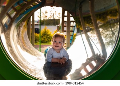 Funny Caucasian Toddler Girl Playing On The Playground, Lies In A Tube. Outdoors. Happy Children's Day.