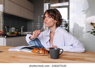 Funny Caucasian Woman With Pencil Near Mount Look Away . Attractive Short Hair Woman Sitting At The Kitchen With Notebook And Breakfast. Concept Of Morning Mood In Home 