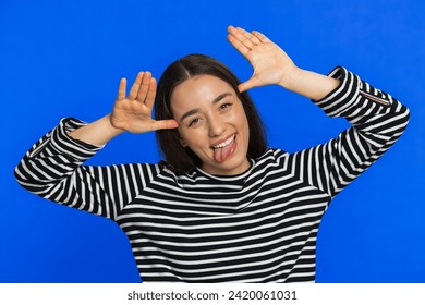 Funny Caucasian woman showing tongue making faces at camera, fooling around, joking, aping with silly face, teasing, bullying, abuse disrespect. Joyful young girl isolated on blue background indoors