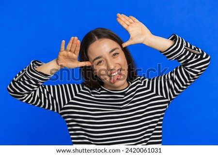 Funny Caucasian woman showing tongue making faces at camera, fooling around, joking, aping with silly face, teasing, bullying, abuse disrespect. Joyful young girl isolated on blue background indoors