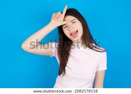 Funny Caucasian woman wearing striped T-shirt over blue background makes loser gesture mocking at someone sticks out tongue making grimace face.
