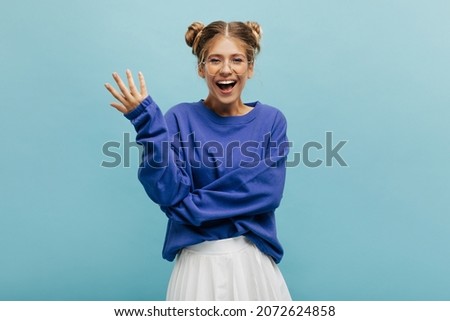 Funny caucasian young girl with two blond tufts of hair on head on blue background. Model in transparent glasses smiles with her mouth open, wearing sweatshirt. Positive emotion concep