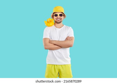 Funny Cheerful Guy Enjoying His Summer Holiday. Studio Portrait Of Happy Young Man In White T Shirt, Yellow Shorts, Bucket Hat And Sun Glasses Standing With Toy Rubber Duck On His Shoulder And Smiling