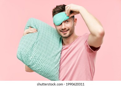 Funny cheerful man in sleeping mask holding pillow in hands looking to camera over pink background. Relax, good morning concept