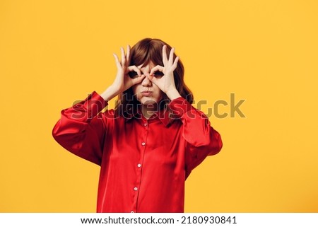 a funny, cheerful woman stands in a stylish red shirt on a yellow background and looking into the camera happily shows the OK sign with her fingers, holding her hands in front of her eyes like a mask