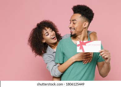 Funny cheerful young african american couple friends woman man in gray green clothes posing hugging hold gift certificate looking at each other isolated on pastel pink color background studio portrait
