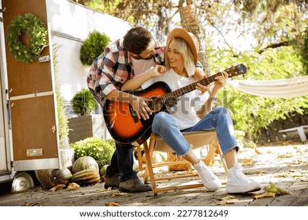 Funny and cheerful young couple. The guy plays the guitar, sing and dance together against the backdrop of a travel trailer. Rest outside the city. Happy people enjoy. Weekends in the summer time.