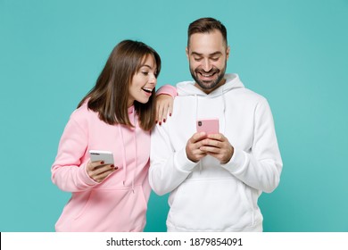 Funny cheerful young couple two friends man woman 20s wearing white pink casual hoodie using mobile cell phone typing sms message isolated on blue turquoise colour wall background studio portrait