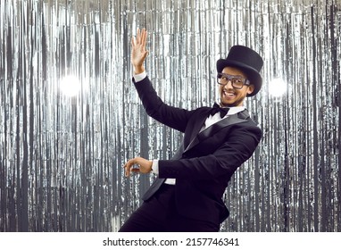 Funny cheerful young guy having fun at party. Happy black man in suit, glasses, bow tie and classic top hat dancing, smiling and waving his hand at camera against shiny silver foil fringe background