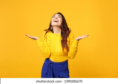 Funny cheerful young woman in sweater, blue trousers looking up spreading pointing hands aside isolated on yellow orange wall background. People sincere emotions lifestyle concept. Mock up copy space