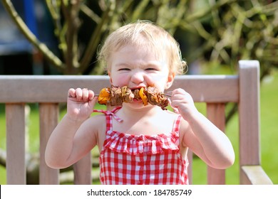 Funny Child, Adorable Blonde Toddler Girl In Red Dress Messy Around Mouth Eating Delicious Meat Made On Bbq Sitting Outdoors In The Garden On A Wooden Chair On A Sunny Summer Day