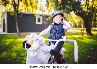 Funny Child Caucasian Girl Blonde In A Bicycle Helmet Near A Purple Bike With A Basket In Outside The Park On A Green Lawn Grass Cart At Home. Plays Uses Hand Phone With Toothy Smile.