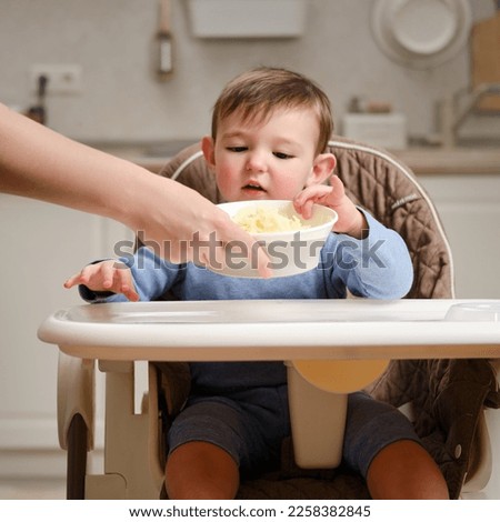 A funny child is eating a grated apple sitting on a kitchen chair. Happy baby boy eat food. Kid aged one year and four months