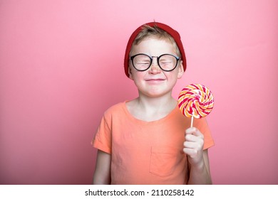 Funny child looking on big lollipop on pink background. Cute eight years boy with close eyes and candy in hand. Happy kid eating sweet food. School pupil.