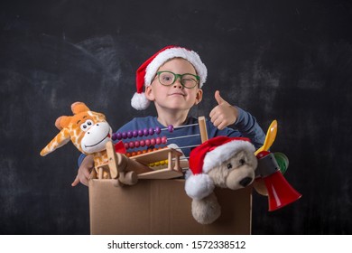 Funny Child In Santa Clause Hat Playing Making Delivery For Kids. Happy School Boy Child Holding Cardboard Box With Toys. Delivery And Donation Concept. Happy Xmas And New Year Holiday! Christmas Gift