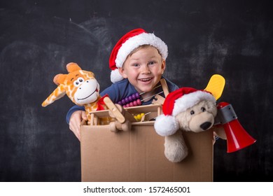 Funny Child In Santa Clause Hat Playing Making Delivery For Kids. Happy School Boy Child Holding Cardboard Box With Toys. Delivery And Donation Concept. Happy Xmas And New Year Holiday! Christmas Gift