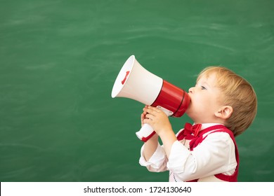 Funny Child Student Speaking By Megaphone In Class. Happy Kid Shouting By Loudspeaker Against Green Chalkboard. Online Education And E-learning Concept. Back To School