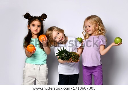 Funny children in colorful casual clothes. They are smiling and holding pineapple, oranges and green apples, posing isolated on white background. Childhood, healthy nutrition. Close up, copy space