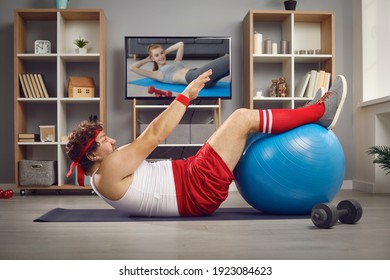 Funny chubby guy doing physical exercise with fitness ball lying on floor at home. Side view of man watching TV video lesson and having sports workout after gaining weight and getting fat in lockdown