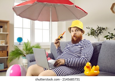Funny chubby man is shocked that he is forced to spend summer vacation at home because of work. Humorous bearded man working on laptop sitting on sofa in living room under beach umbrella.
