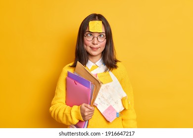Funny clever female nerd crosses eyes has sticker with drawn graphic on forehead prepares research work studies from home holds folders isolated over vivid yellow background. Paperwork concept