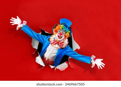 Funny clown with open arms  looking through the ripped paper hole, man entertainer as Joker in a suit and wig, with clown whiteface makeup. Trickster, jester, professional actor at events