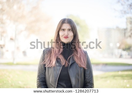 Funny comic girl crosses eyes, puts lips, makes grimace, foolishes after all day studying. Clueless young woman nerd with awkward expression has fun alone, plays fool. Wears biker jacket.